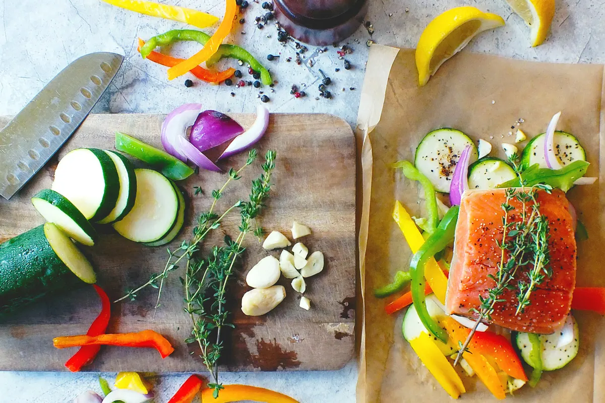 Bright orange salmon arranged on a sheet of parchment paper, surrounded by sliced zucchini, bell peppers and onions next to a cutting board with more freshly sliced veggies, garlic and herbs.