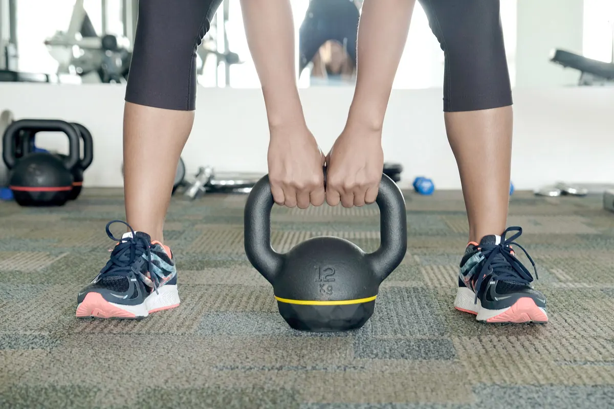 A woman in athletic clothes bending over to pick up a kettlebell.