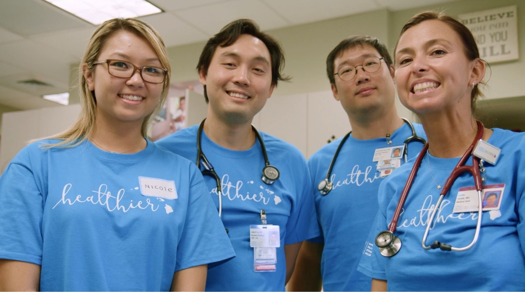 Group of healthcare workers in matching blue shirts and stethoscopes stand together, smiling at the camera.