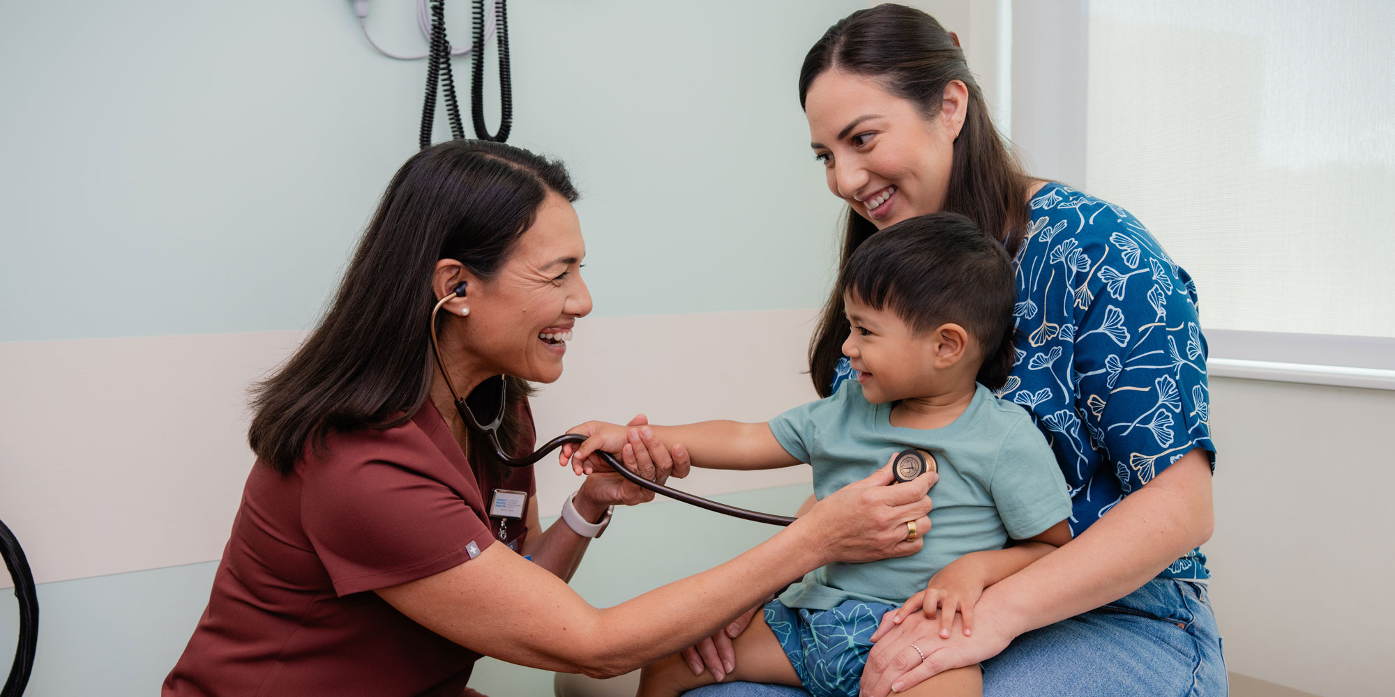 a happy healthcare provider smiles at a child while checking his heartbeat in a clinic exam room as his mother holds him and smiles at the healthcare worker.