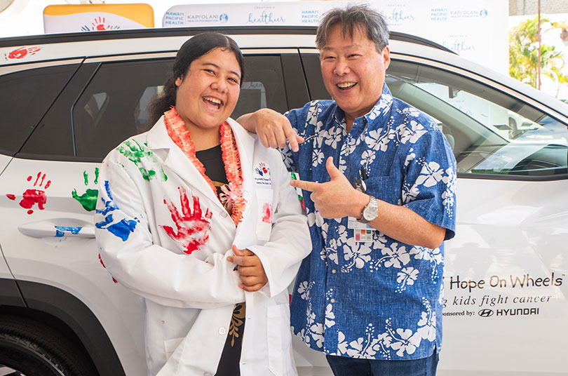 Two people smiling in front of a hope on wheels vehicle covered in green, blue, and red painted handprints.