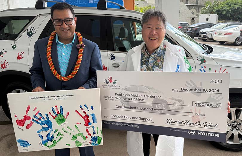 Two people in front of a Hyundai, one holding a sign with children's handprints, the other in a white coat holding a giant check for $100,000.