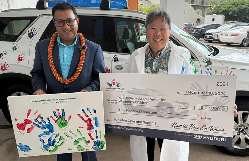 Two people in front of a Hyundai, one holding a sign with children's handprints, the other in a white coat holding a giant check for $100,000.