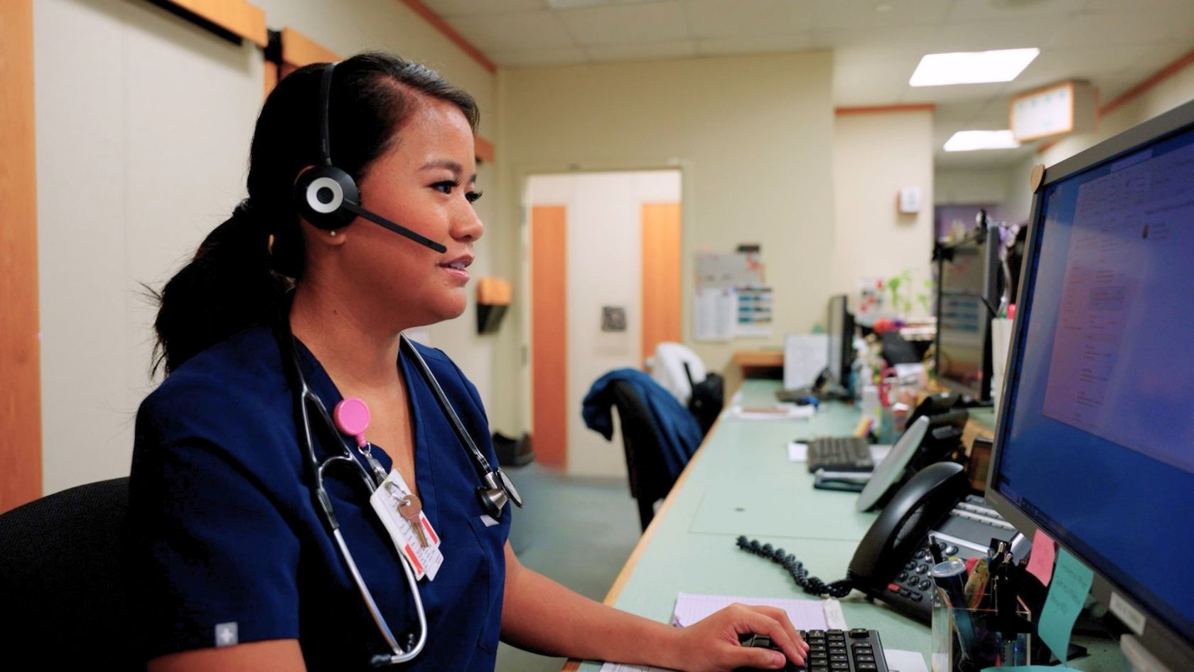 Healthcare worker wearing a headset and stethoscope enters information on a computer at a nurses’ station.