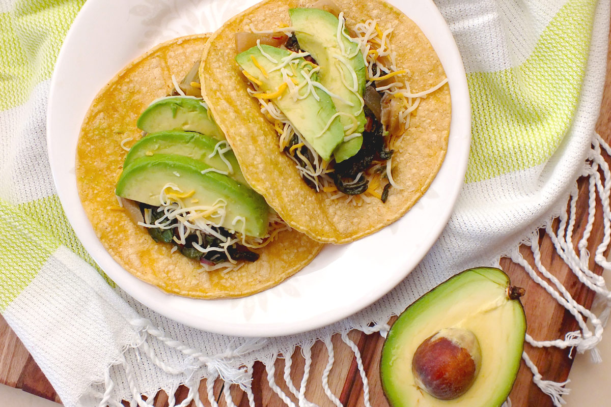 Chard, avocado, and cheese sit on corn tortillas on a white plate.