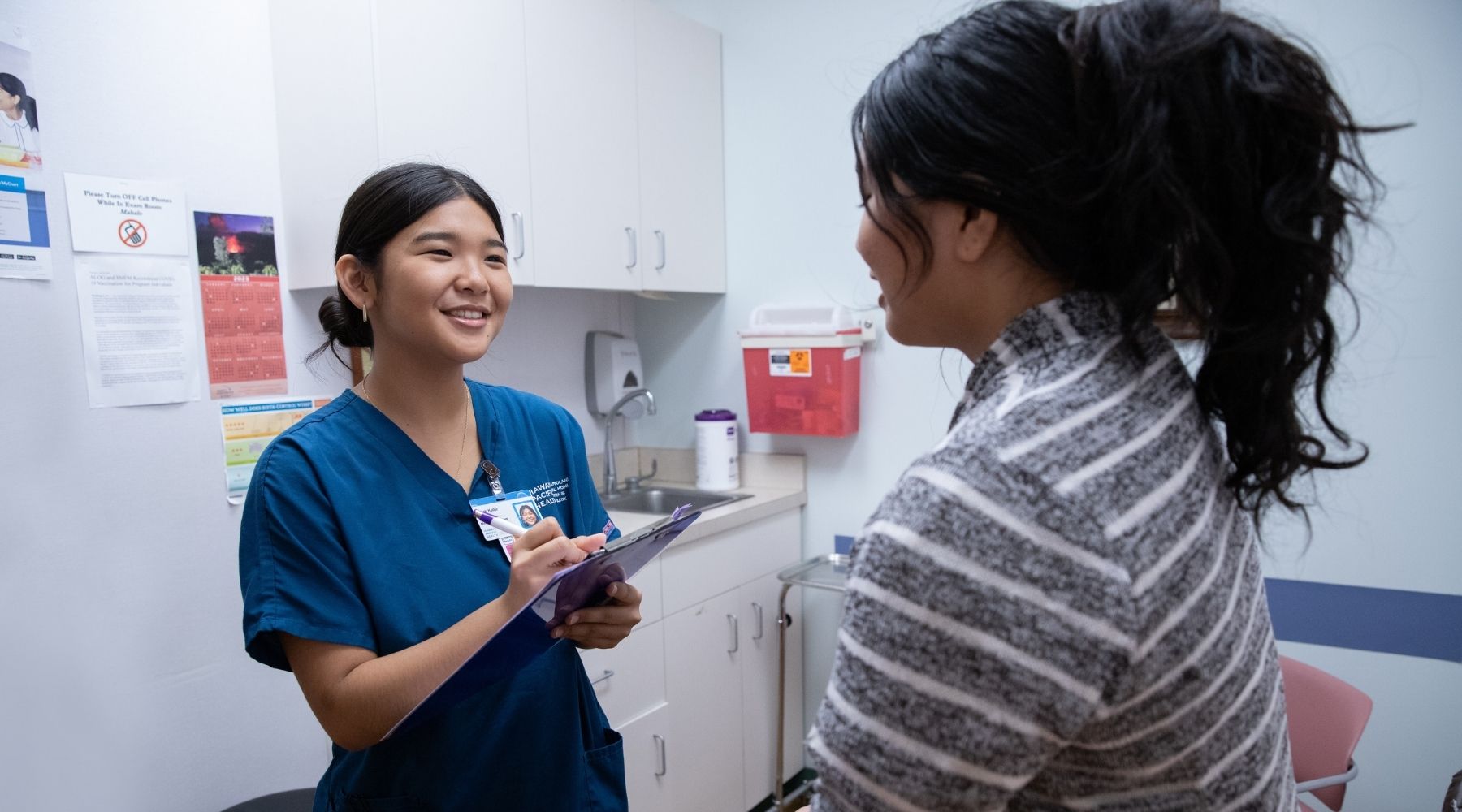 Healthcare worker takes notes on a clipboard while speaking with a patient in an exam room.