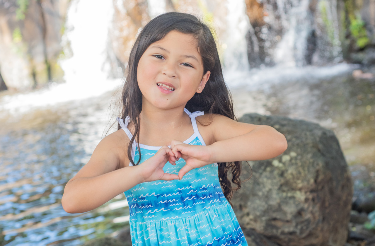 Leila-Rose Coloma makes a heart symbol with her hands while standing in front of a waterfall