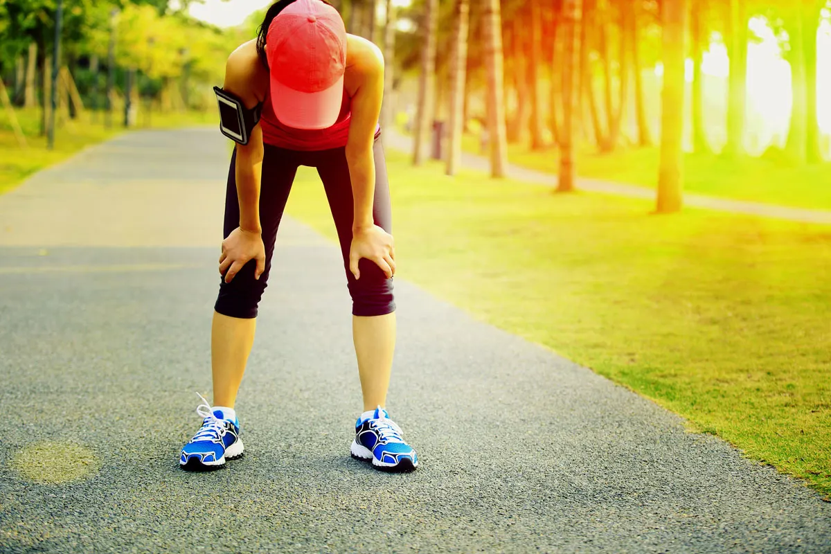 A young athletic woman bent over with hands on knees as sun beats down on her back.