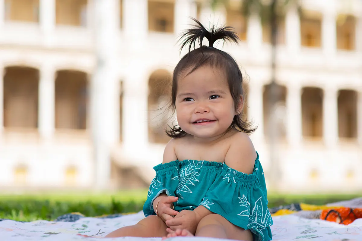 A 9-month-old girl sits on a colorful blanket in front of Iolani Palace and smiles at the camera.