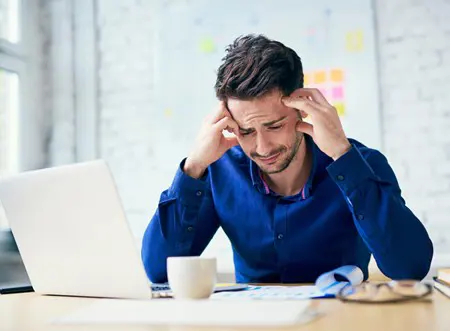 A man sits in front of a laptop, head in hands, looking stressed.