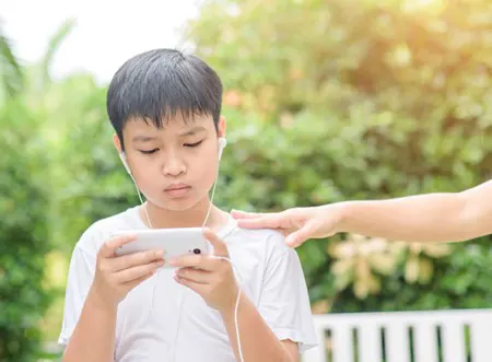 A kid is watching a phone's screen with headphones in while some one reaches out to touch their shoulder.