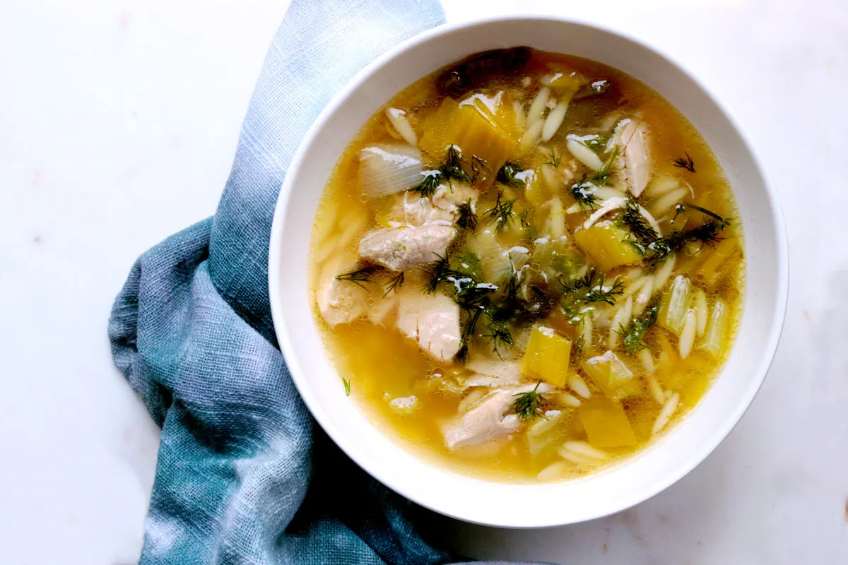 Golden broth, rich soup and green garnish sit in a white bowl with a blue napkin on a white table.