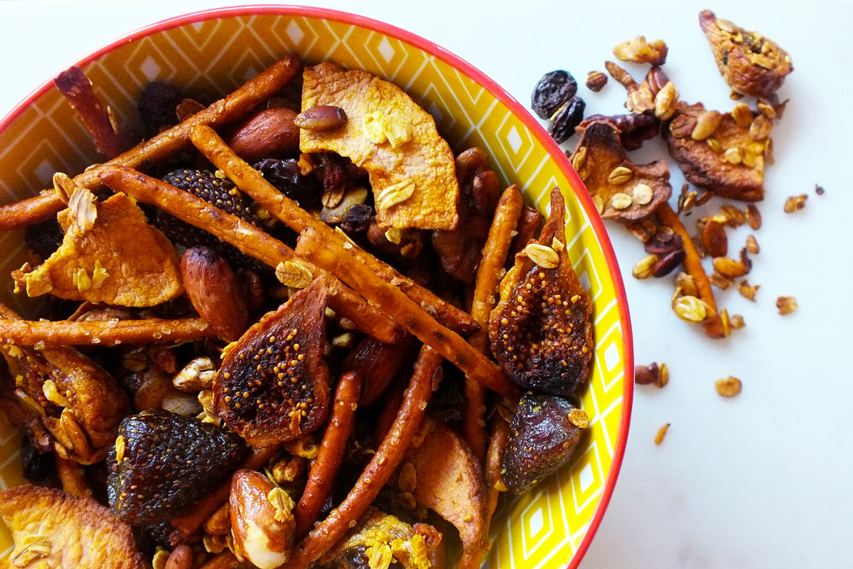 A yellow bowl sits on a marble counter top filled to the brim with Sweet & Salty Crunch Mix, with some extra mix overflowing onto the table top.