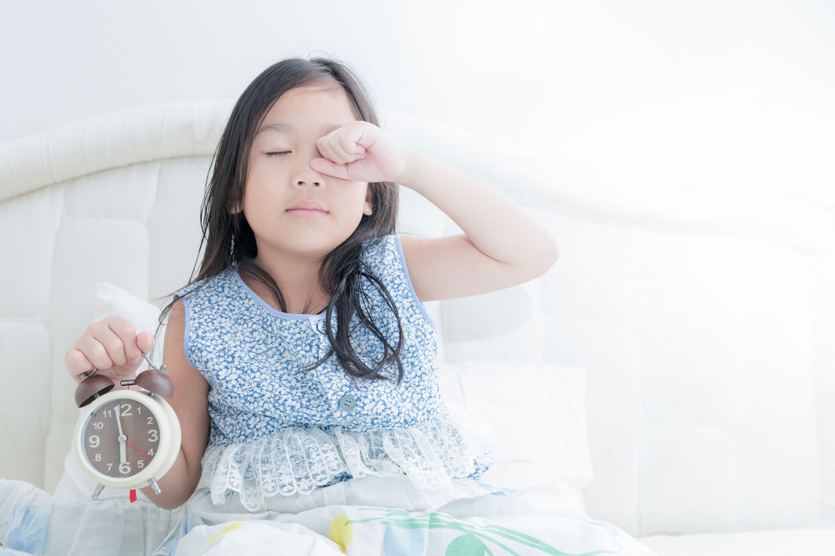 A young girl sitting up in bed holding an alarm clock and rubbing sleep from her eyes. 