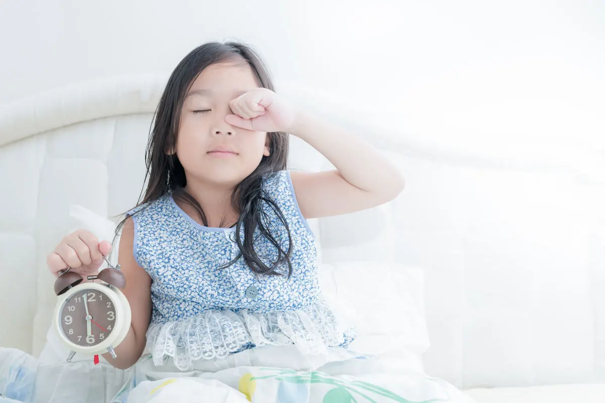 A young girl sitting up in bed holding an alarm clock and rubbing sleep from her eyes. 