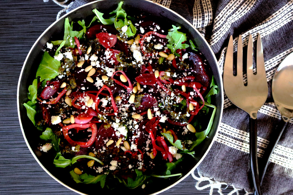 A bowl of Roasted Beet Salad with Basil-Lemon Pesto sits on a dark counter top next to a napkin and serving utensils.