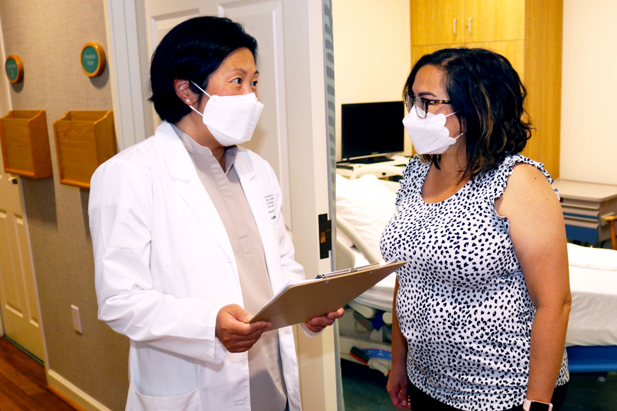 Physician in a white coat, holding a clipboard, and speaking with a nurse practitioner. Both are wearing masks.