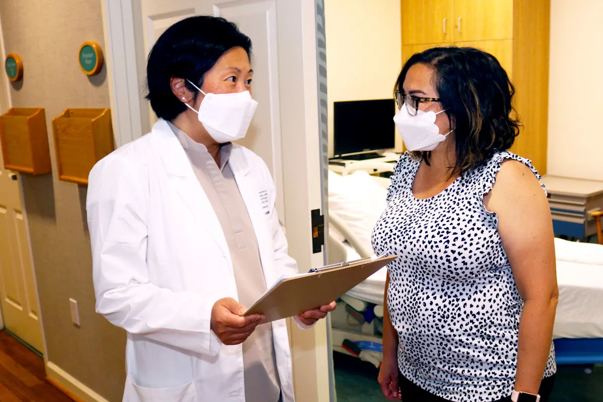 Physician in a white coat, holding a clipboard, and speaking with a nurse practitioner. Both are wearing masks.