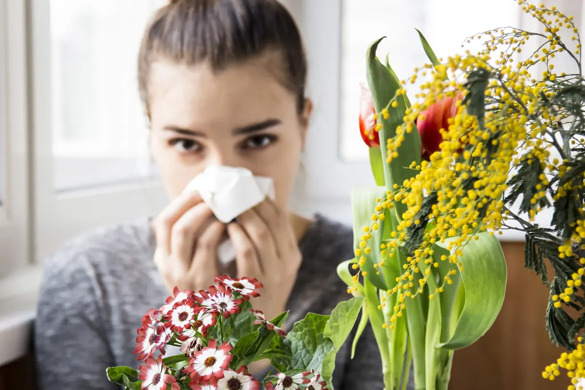 A woman blowing her nose into a tissue in front of a bouquet of flowers.