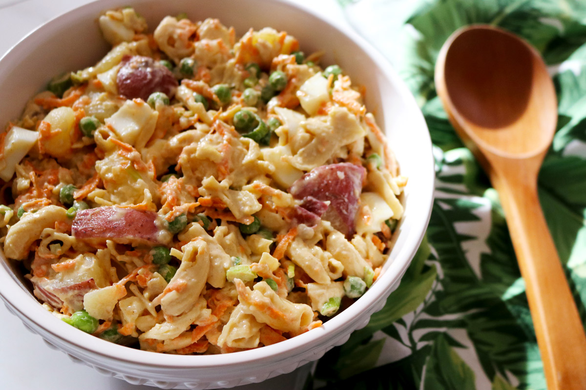 A bowl of Healthier Hawaiian-Style Potato-Mac Salad in a porcelain salad bowl next to a palm-print napkin and wooden serving spoon.