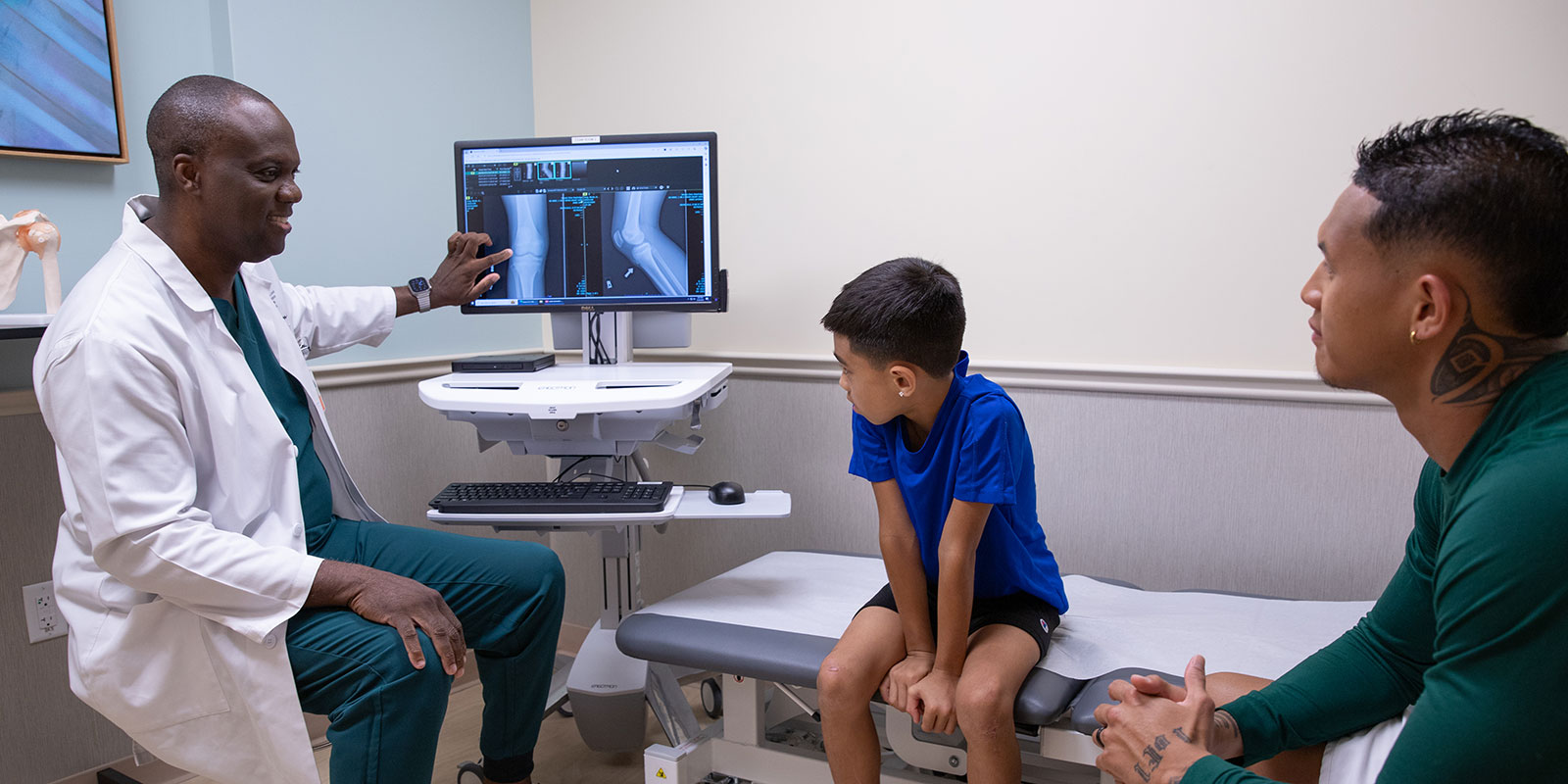 Doctor pointing at xray image on screen showing a knee while young patient and his father listen.