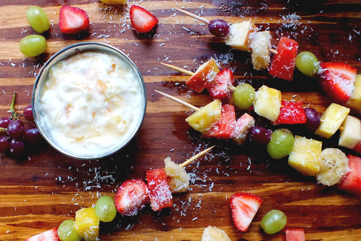 Fresh Fruit Kebabs with Coconut Yogurt Dip sit on a wood cutting board with coconut shavings over top.