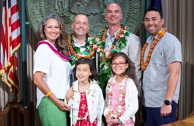 Group shot of family with young children with governor and trust leader in front of Hawaiʻi state seal.