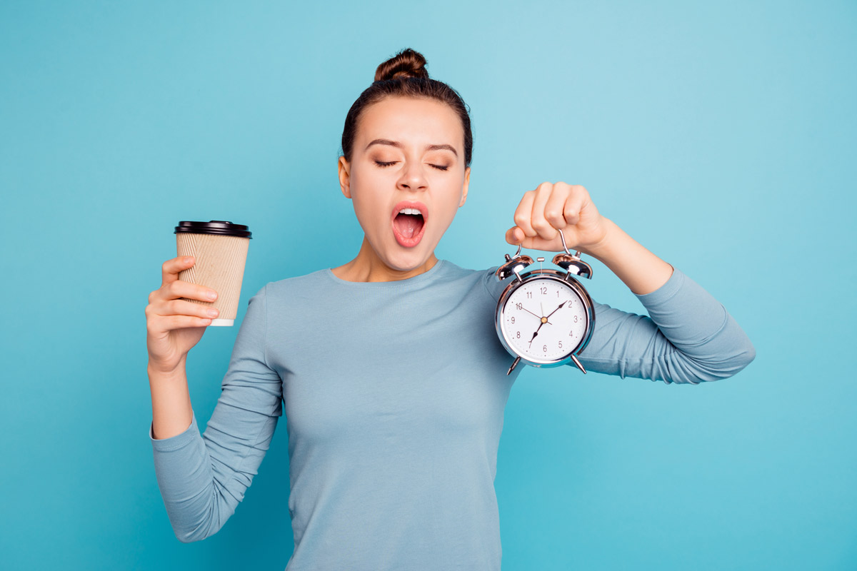A woman yawning holding an alarm clock in one hand and a cup of coffee in the other hand.