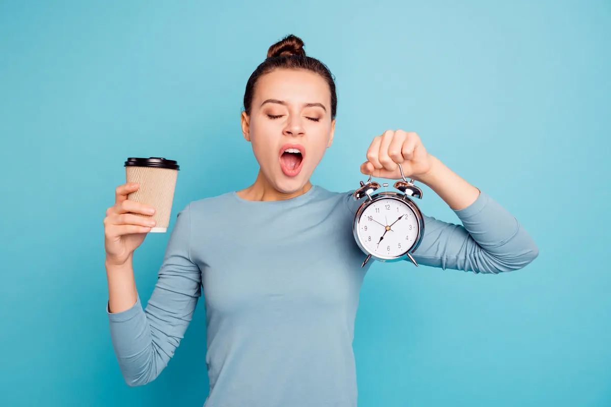 A woman yawning holding an alarm clock in one hand and a cup of coffee in the other hand.