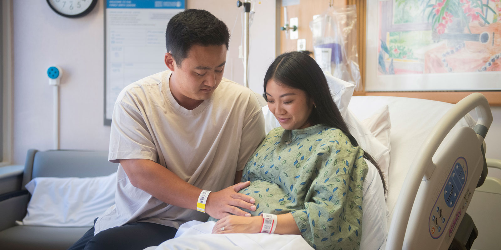 Very pregnant woman in a hospital birth suite with her partner who has a hand on her belly as both gaze down lovingly.