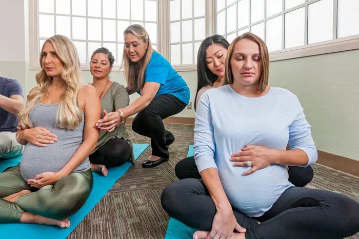 Pregnant women seated on yoga mats as their partners massage their neck and shoulders from behind and an instructor guides one partner.