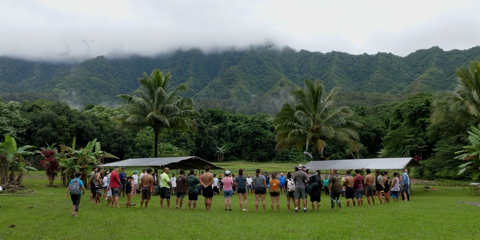 Circle of people in a vibrant green outdoor area with green mountains in the background and dramatic low hanging clouds.