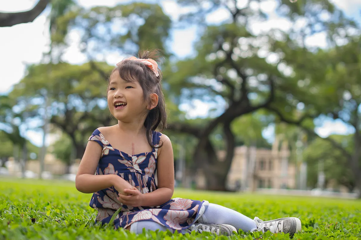 Kiryn smiles while sitting in the grass with trees behind her.