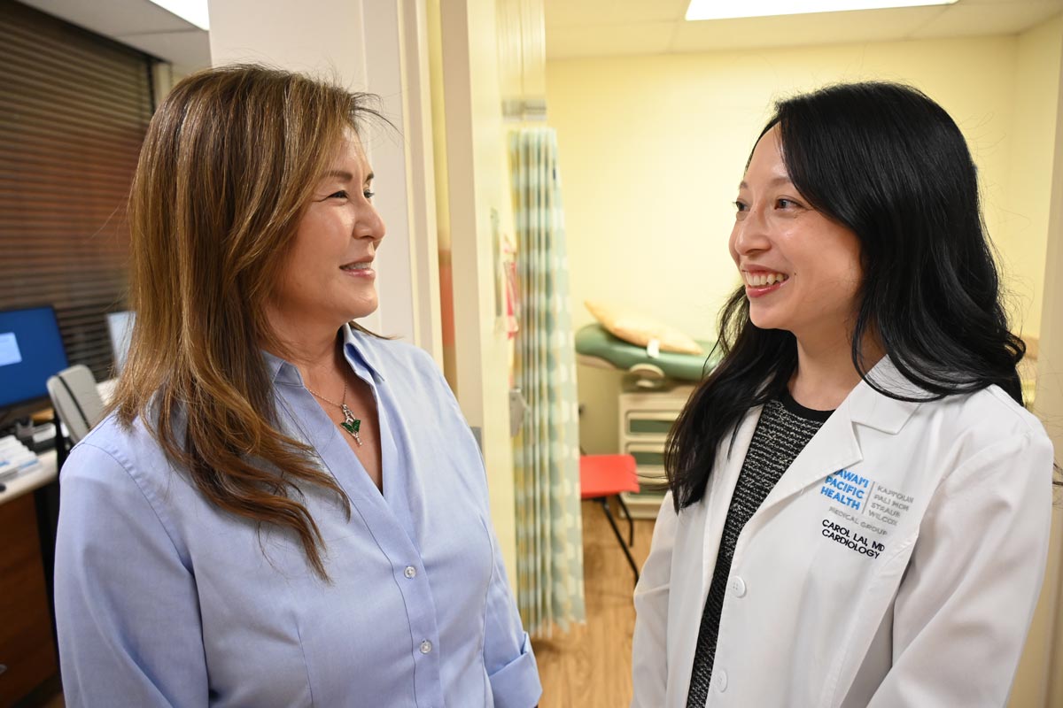 A female doctor and nurse talk to each other in front of an exam room in a clinic.