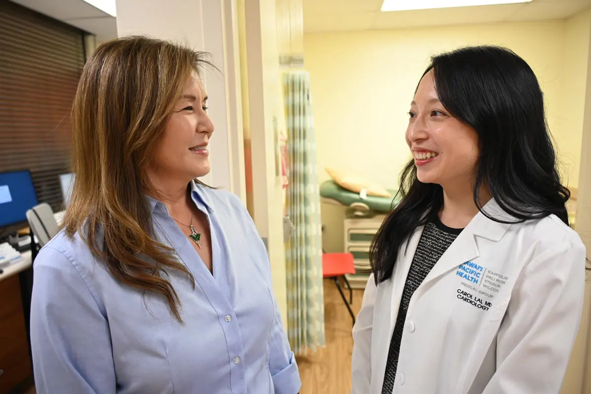A female doctor and nurse talk to each other in front of an exam room in a clinic.