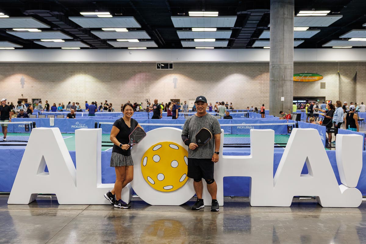 Two pickleball players stand in front of a giant ALOHA sign holding paddles.