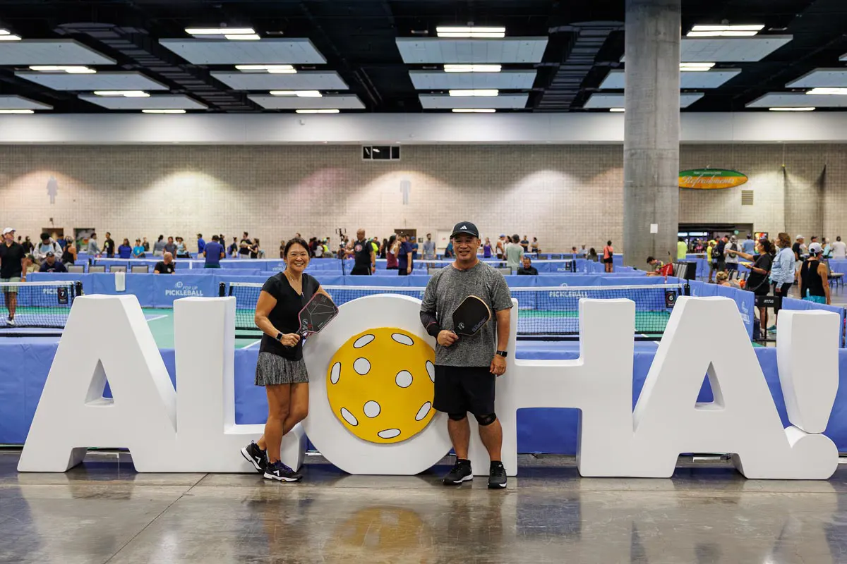 Two pickleball players stand in front of a giant ALOHA sign holding paddles.