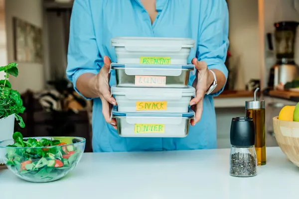 a woman stands in her kitchen and holds up Tupperware labeled for different meals