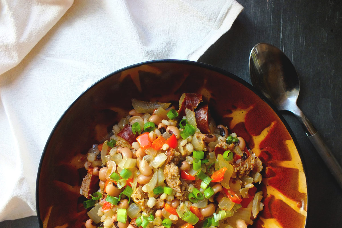 A colorful mix of vegetables, grain and sausage sits in a red and orange bowl with a spoon and napkin nearby,