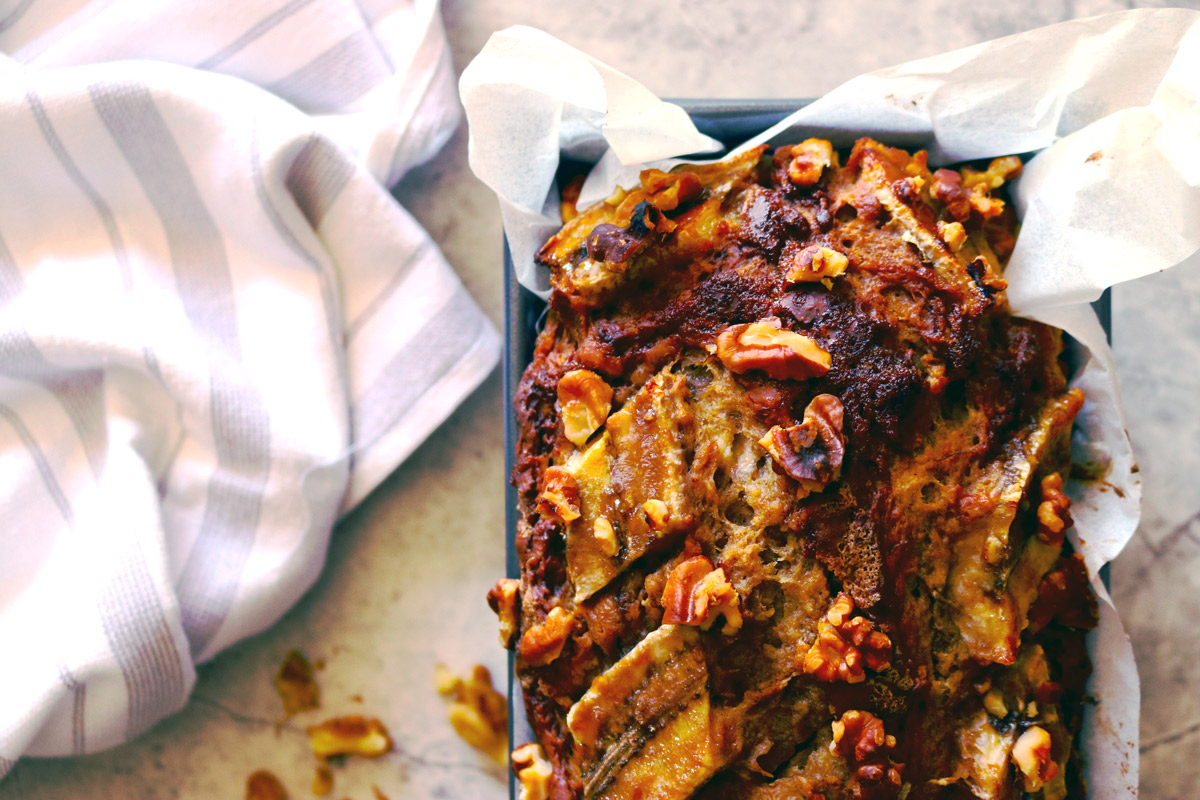 A freshly baked loaf of Banana Walnut Bread in a baking pan cools on a marble counter with a striped dish towel and walnut pieces surrounding the loaf.