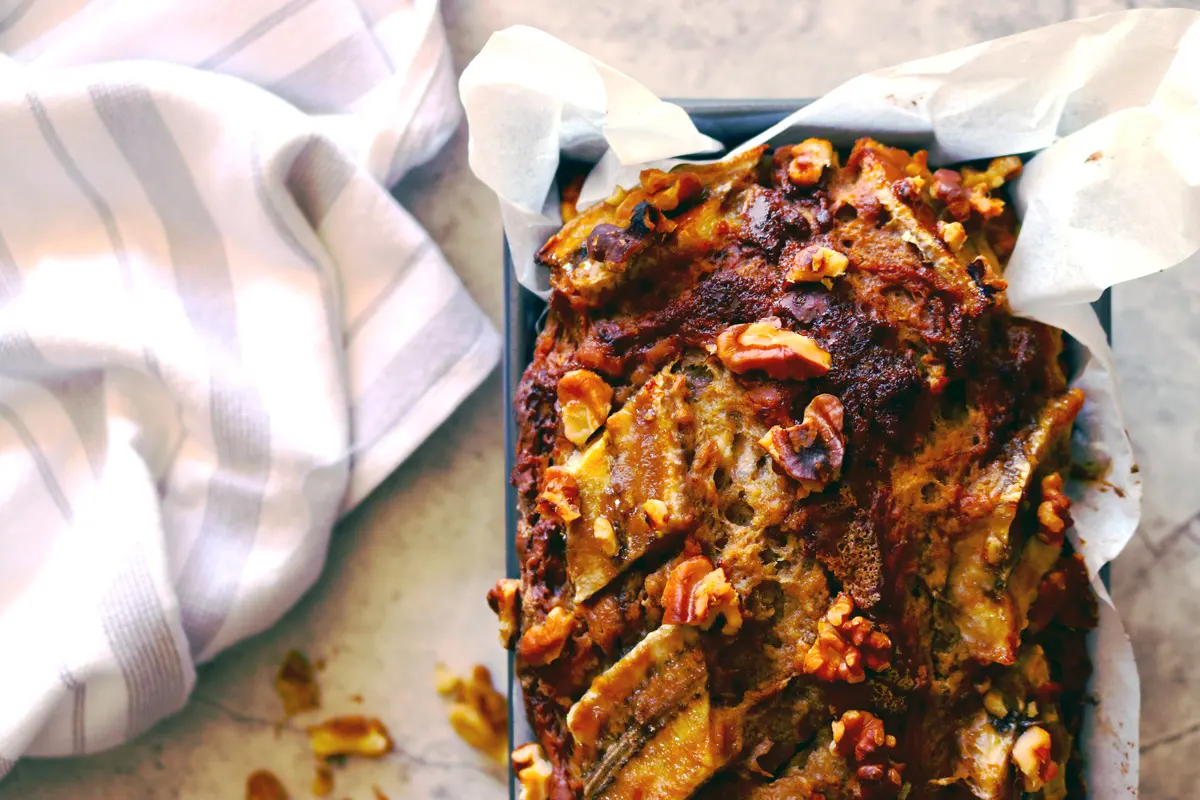 A freshly baked loaf of Banana Walnut Bread in a baking pan cools on a marble counter with a striped dish towel and walnut pieces surrounding the loaf.