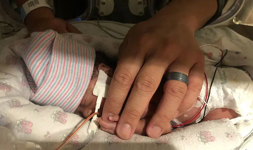 Kalley-Mae as a micropreemie baby resting and holding on to the fingertip of a hand that nearly covers her entire body. 