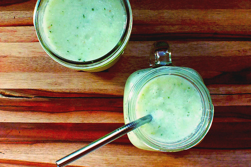 Two glasses of pale green smoothie sit on a wooden board, one with a metal straw.