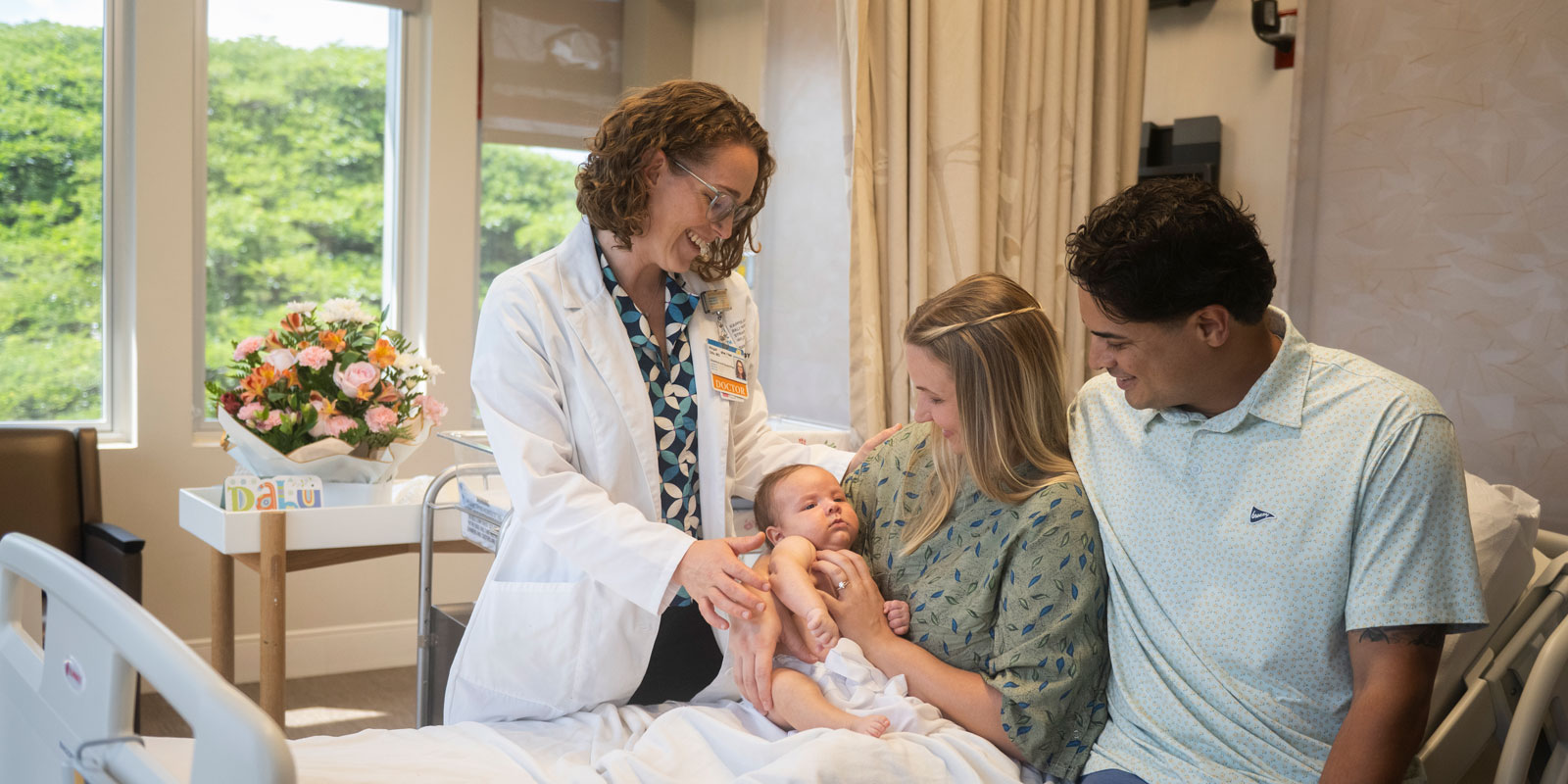 A doctor stands and smiles at a mother, father, and their newborn baby as they sit on a bed in a light-filled room in the Mother Baby Care Unit at Kapiolani Medical Center for Women & Children.
