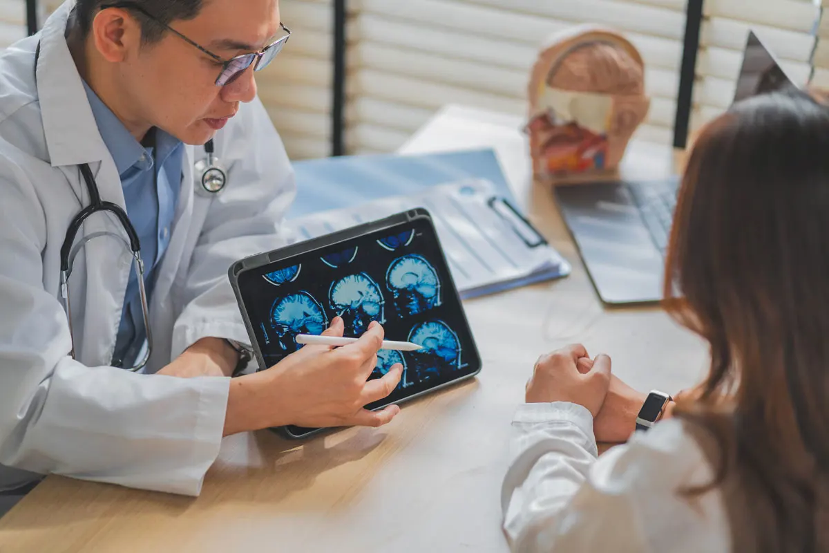 a neurologist sits behind his desk in his office and shows a patient a brain scan on an iPad