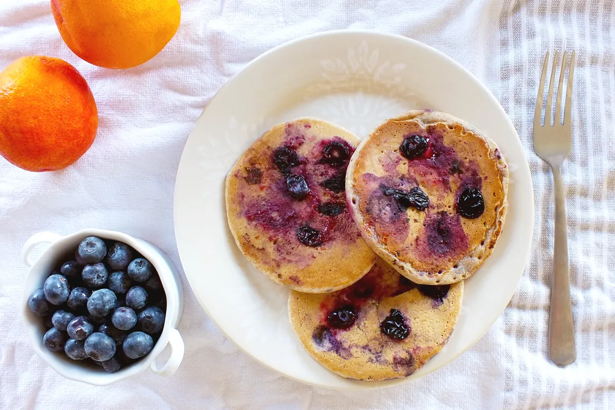 Three blueberry pancakes sit on a white plate with a fork, orange and blueberries next to it.