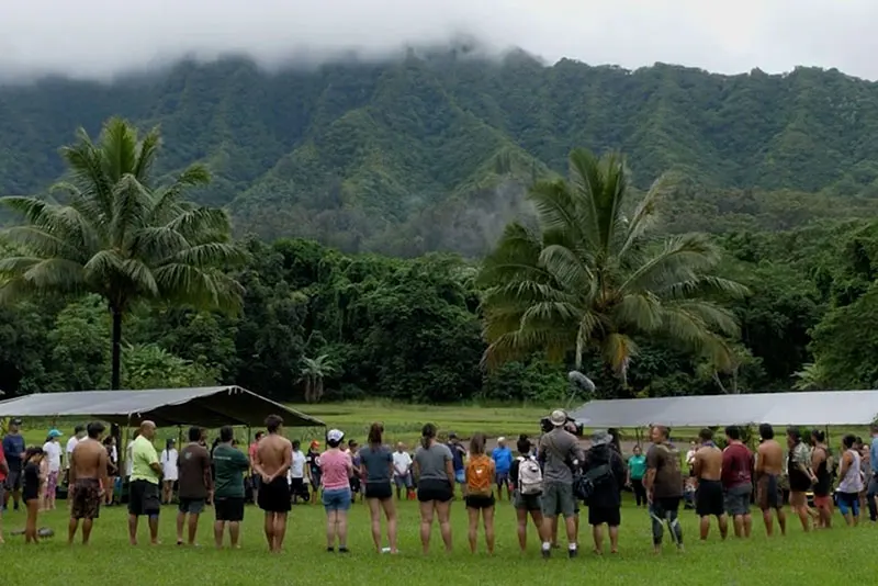 Large group of people standing in a circle in a lush green valley with dramatic green mountains and cloud cover in background.