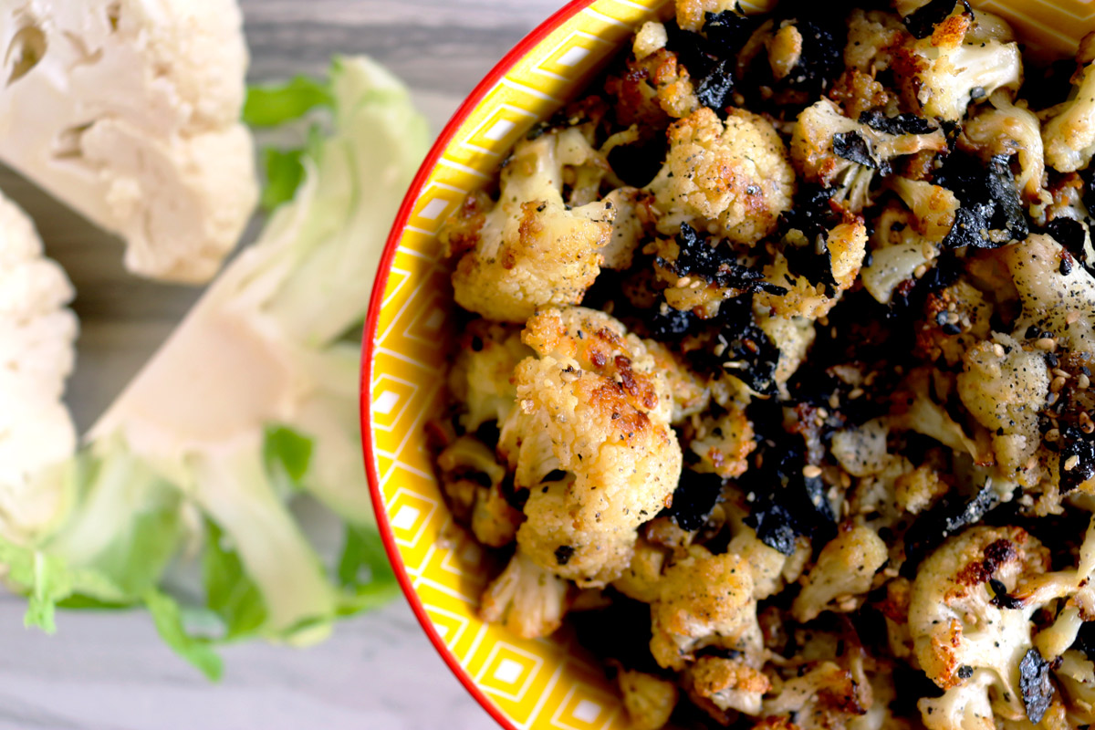 A closeup of a bowl of Cauliflower Hurricane Popcorn on a counter top with cauliflower florets next to it.