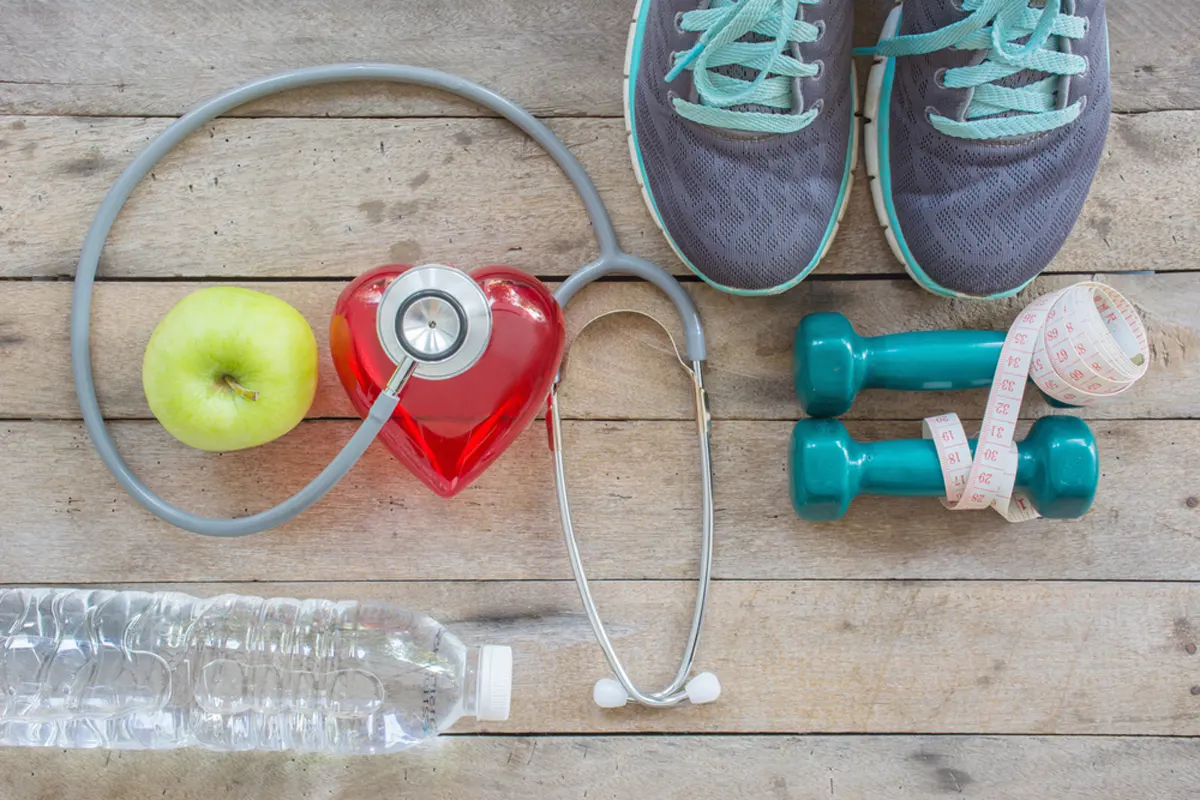Running shoes, weights, a tape measure, an apple and a bottle of water surround a stethescope on a plastic heart.