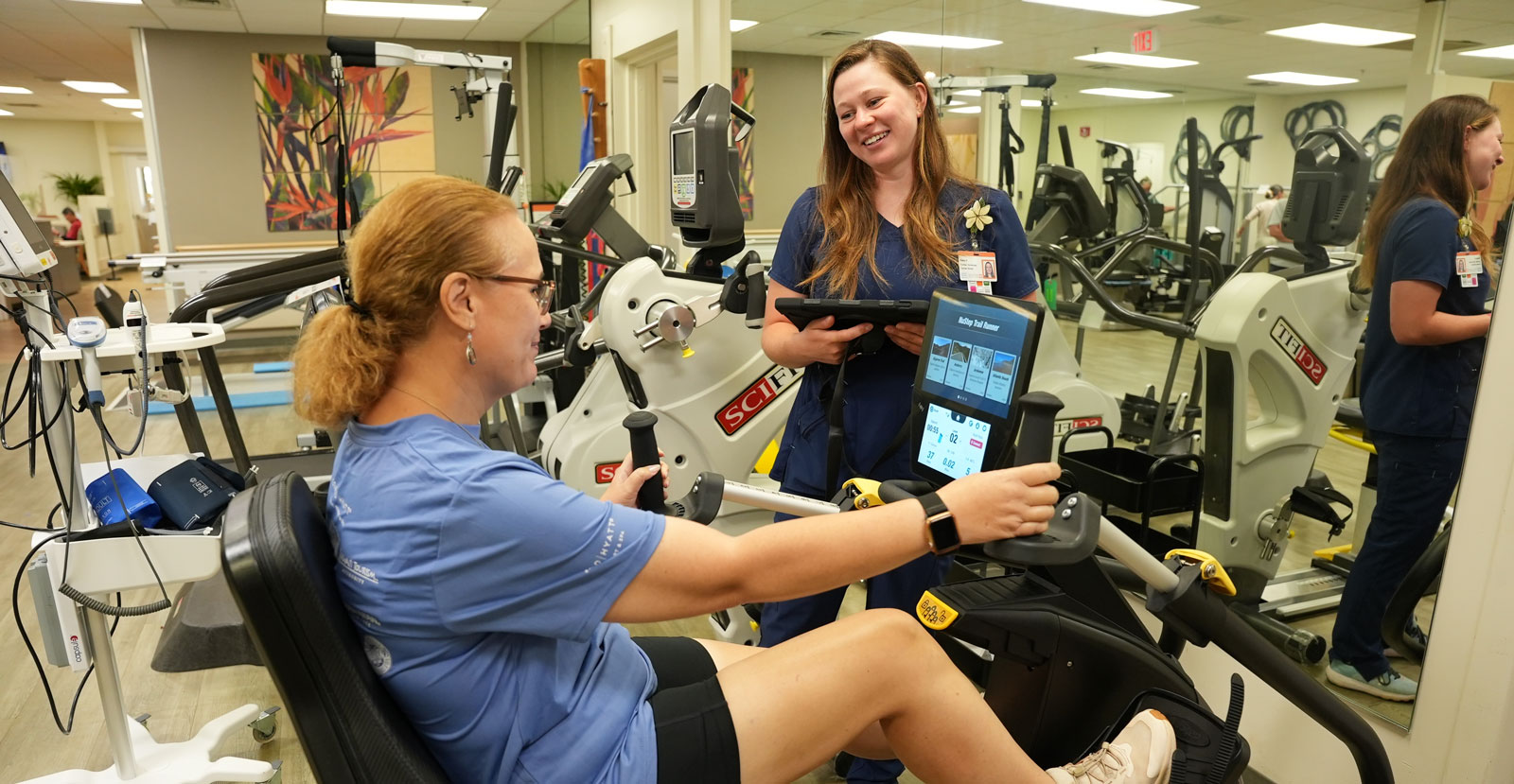 a female patient performs a heart stress test on a stationary bike as a healthcare professional watches and smiles in encouragement in the Wilcox Cardiac Rehab room.